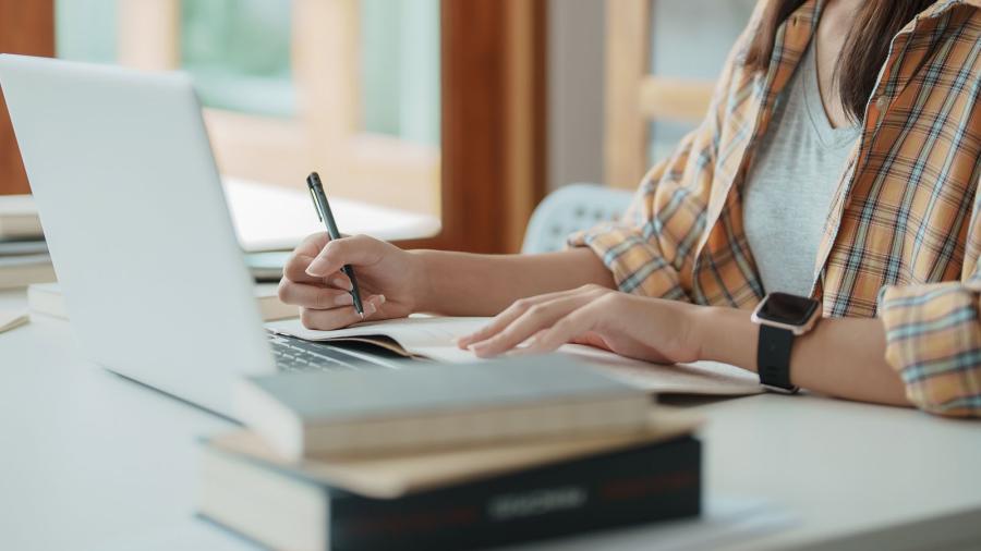 A laptop sits on table while a woman takes notes. 