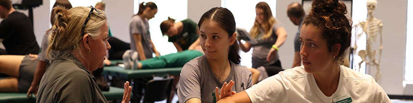 A professor watches as a student practices a technique on another student during the Occupational Therapy lab immersion.